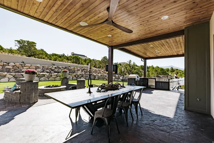 Covered patio with a wood-plank ceiling, ceiling fan, built-in outdoor kitchen, stamped concrete flooring, and a stone retaining wall