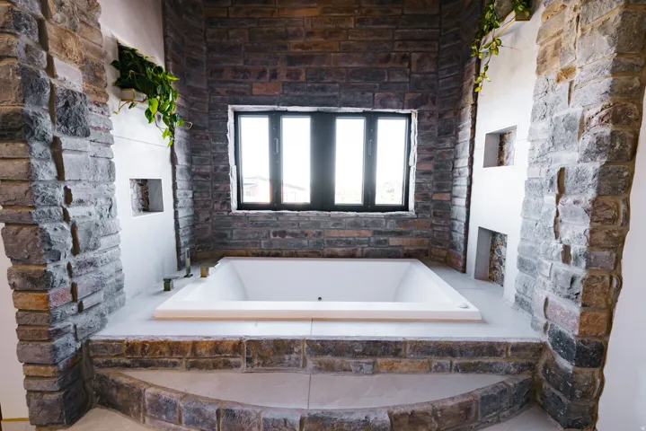 Integrated soaking tub with stone surround, complemented by a large window with dark trim and stone wall details