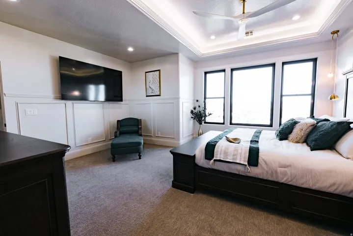 Tray ceiling with recessed lighting and a ceiling fan, three large windows, wainscoting wall detail, and wall-mounted light fixtures