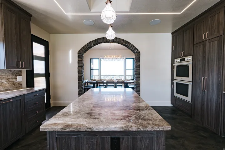 Kitchen featuring a large island with a substantial countertop, dark wood-finish cabinetry, stainless steel double ovens, and a stone archway