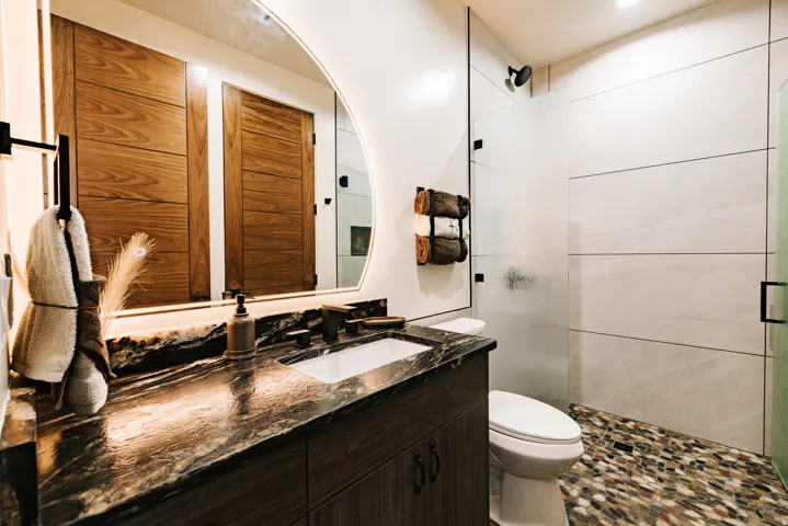Contemporary bathroom featuring a dark wood-finish vanity with a stone countertop, a shower with horizontal wall tiling and a pebble-finish floor, and a frameless oval mirror with integrated backlighting