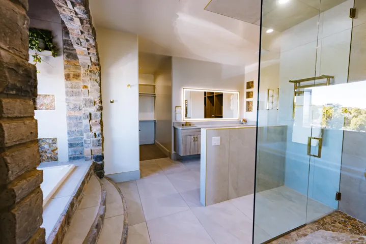 Stone-clad archway leading to a built-in tub, tiled flooring, glass-enclosed shower, single vanity with integrated lighting, and a walk-in closet