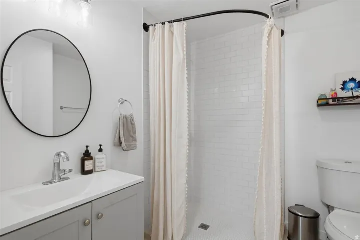 White subway tile shower surround with a mosaic tile floor, vanity with a rectangular sink, wall-mounted circular mirror, and chrome faucet