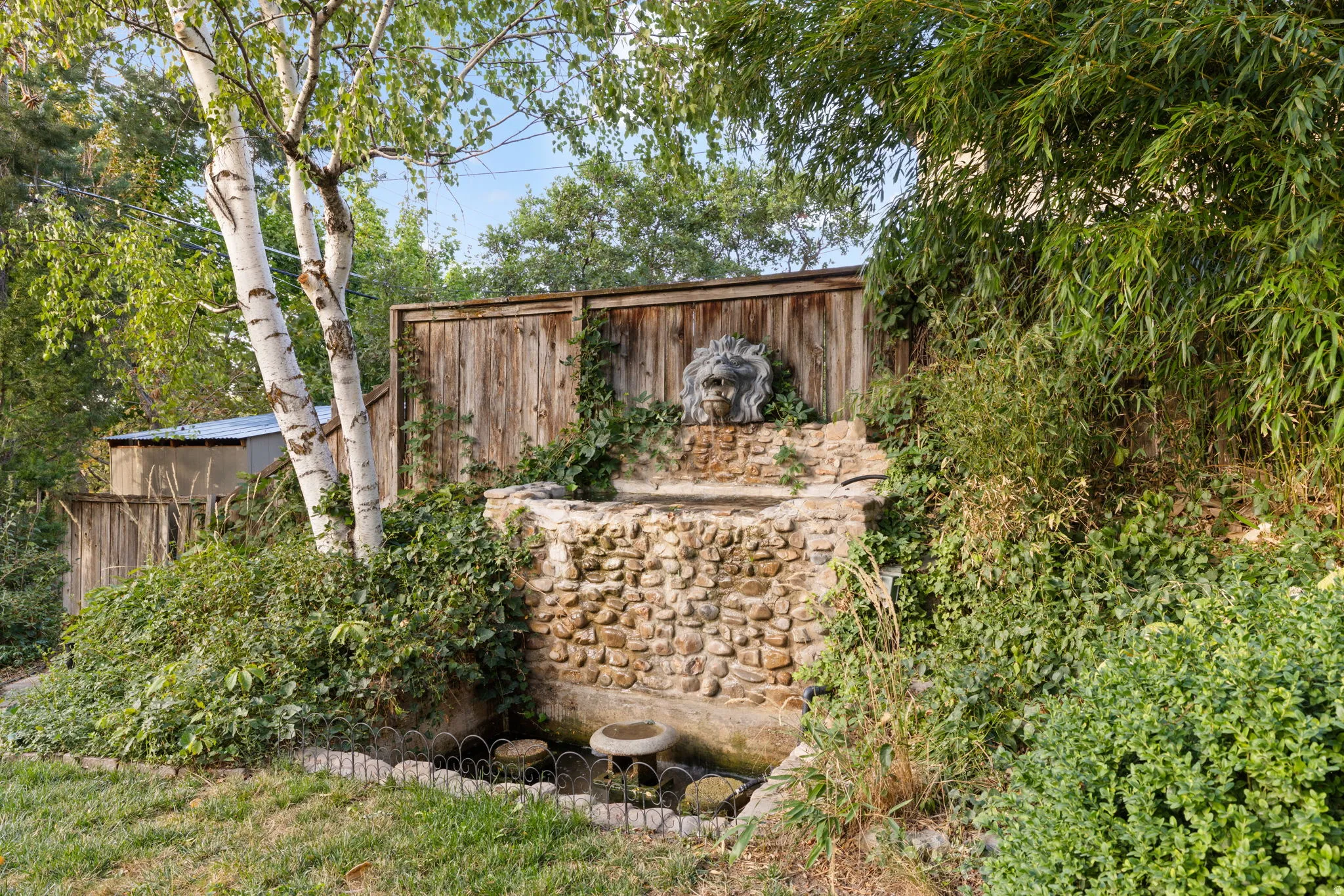 Stone-clad water feature with a lion's head spout, surrounded by mature greenery and a wood privacy fence
