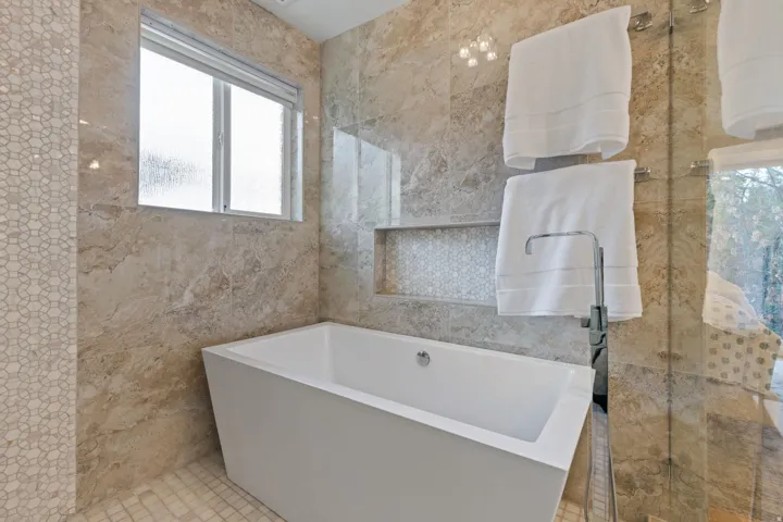 Freestanding soaking tub with a floor-mounted faucet, complemented by full-height natural stone wall tiling, a mosaic tile accent, and a privacy window