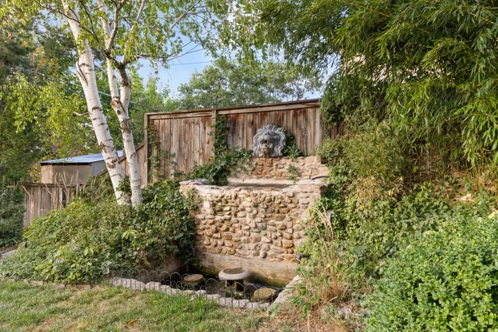 Stone-clad water feature with a lion's head spout, surrounded by mature greenery and a wood privacy fence
