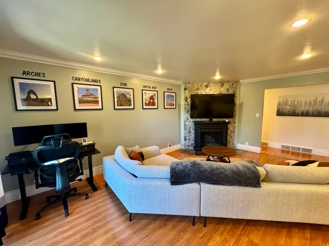 Recessed lighting illuminating the living space, a stone-clad fireplace with a black mantel, wood-finish flooring, and crown molding