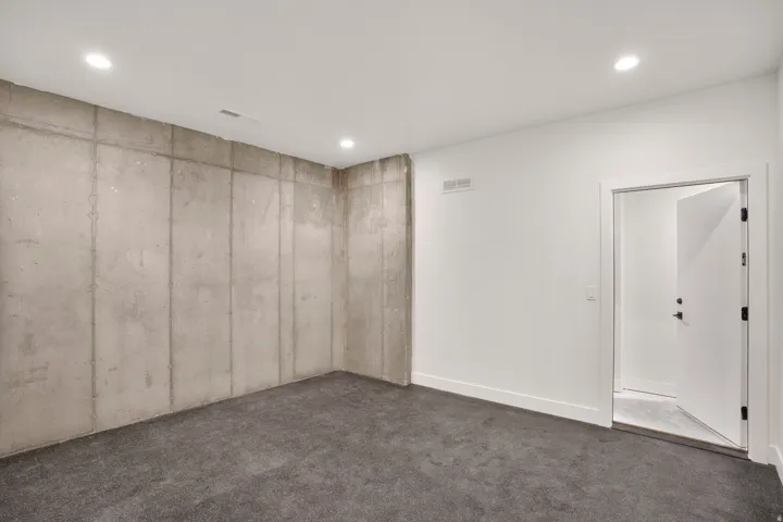 Recessed lighting, concrete wall, white painted walls, gray carpeting, interior door with dark hardware