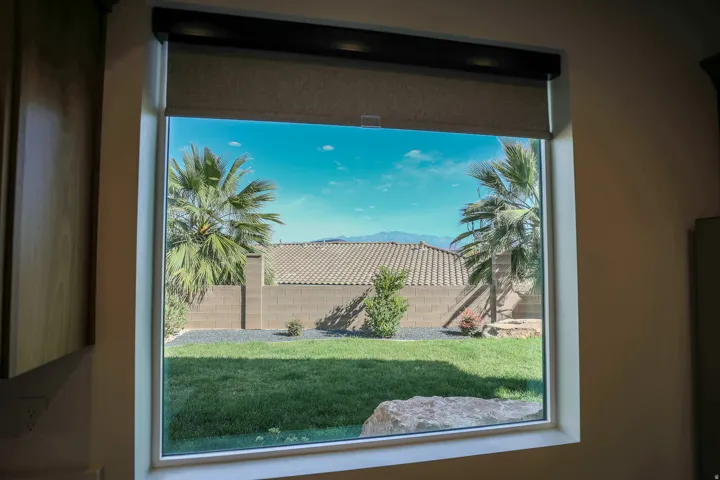 ADU - Window with roller shades overlooking a yard with manicured lawn, landscaping rocks, and palm trees