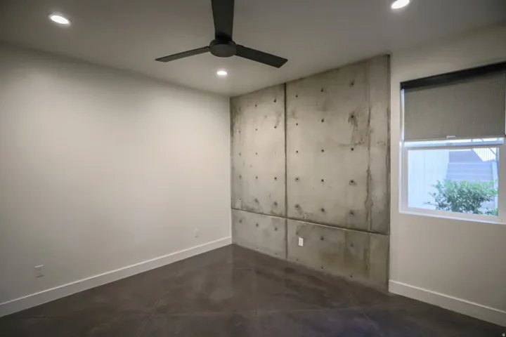 ADU - Exposed concrete accent wall, recessed lighting, ceiling fan, dark concrete flooring, and a window with a roller shade
