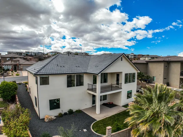 Aerial view - contemporary stucco exterior with a tiled roof, featuring an upper-level balcony with metal railings, a lower-level patio, and landscaped grounds with gravel and mature plantings
