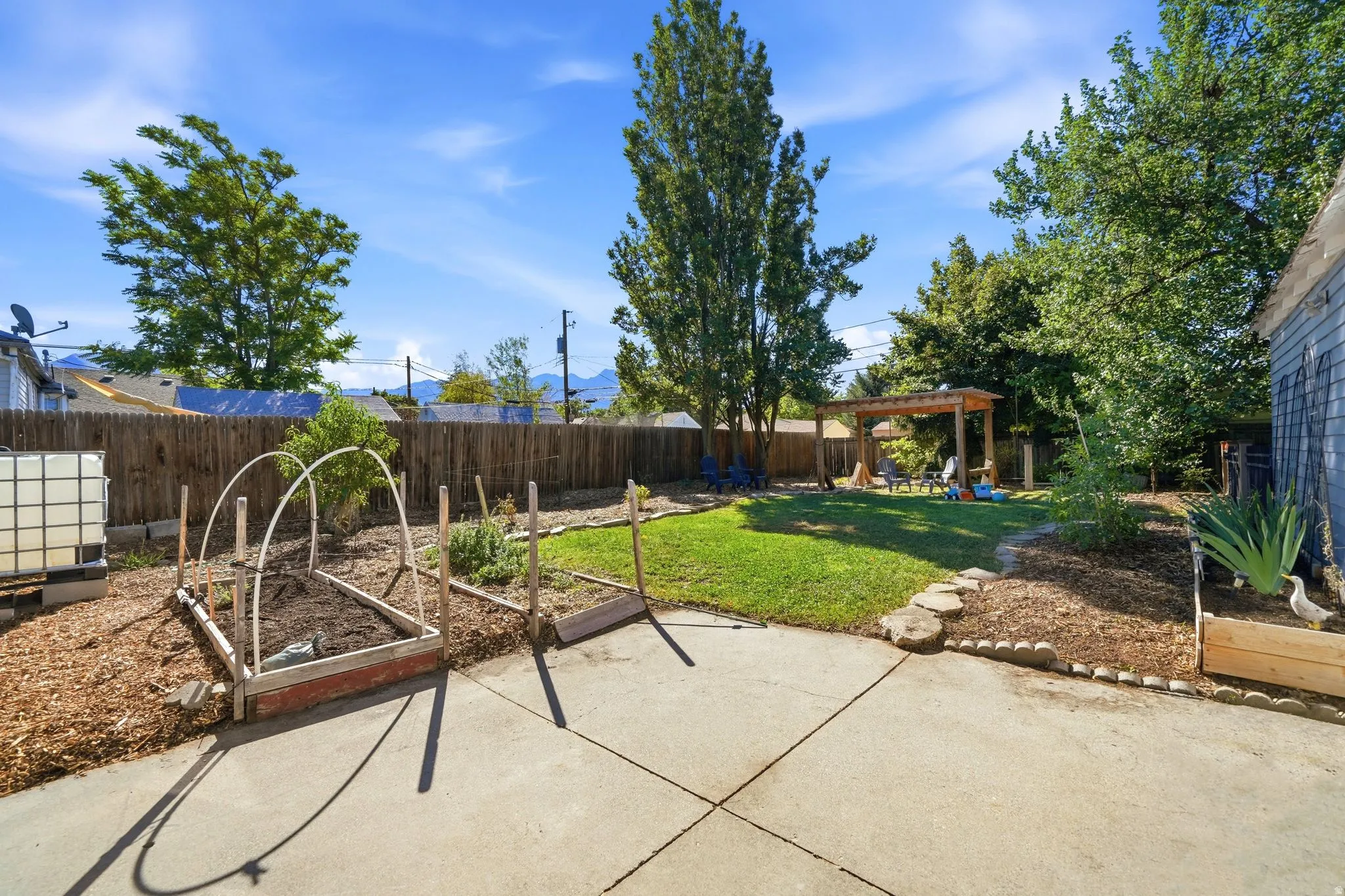 Expansive backyard featuring a concrete patio, a manicured lawn, a wooden privacy fence, mature trees, and a wooden pergola structure