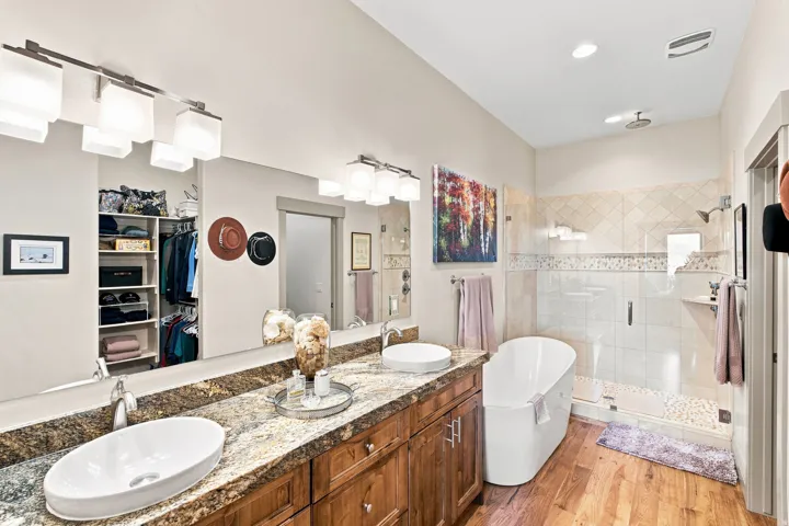 Dual vessel sinks set in a granite-finish countertop, wood-finish cabinetry, a freestanding soaking tub, a glass-enclosed shower, and wood-finish flooring