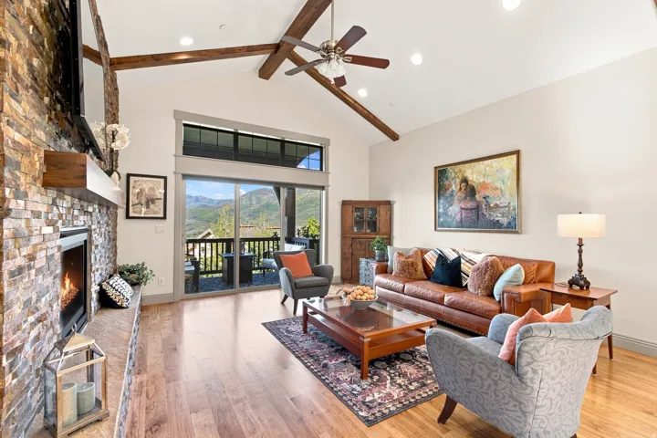 Vaulted ceilings with exposed wood beams, a stone-clad fireplace, wood-finish flooring, and a sliding glass door leading to an outdoor deck