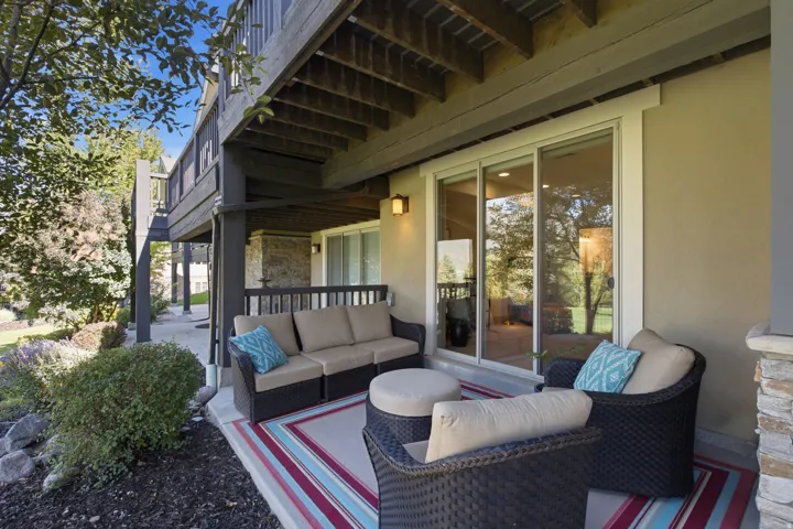 Ground-level patio with concrete flooring, covered by an elevated deck, featuring an exterior wall light and sliding glass doors