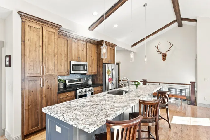 Kitchen featuring a vaulted ceiling with exposed wood beams, stainless steel appliances, wood-finish cabinetry, a large kitchen island with a built-in sink, and a tile backsplash