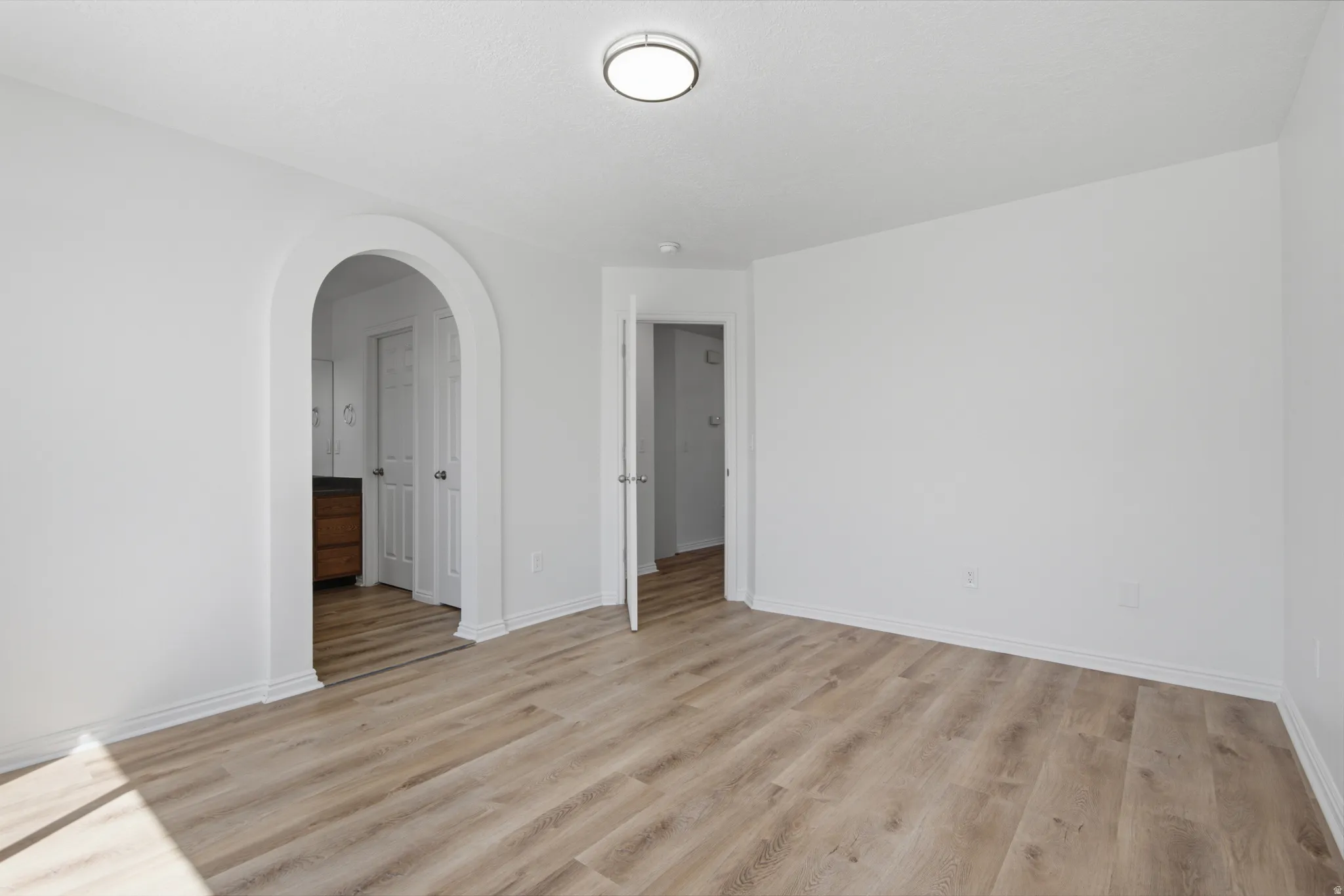Wood-finish flooring throughout, bright white walls, contemporary ceiling light fixture, and an arched doorway