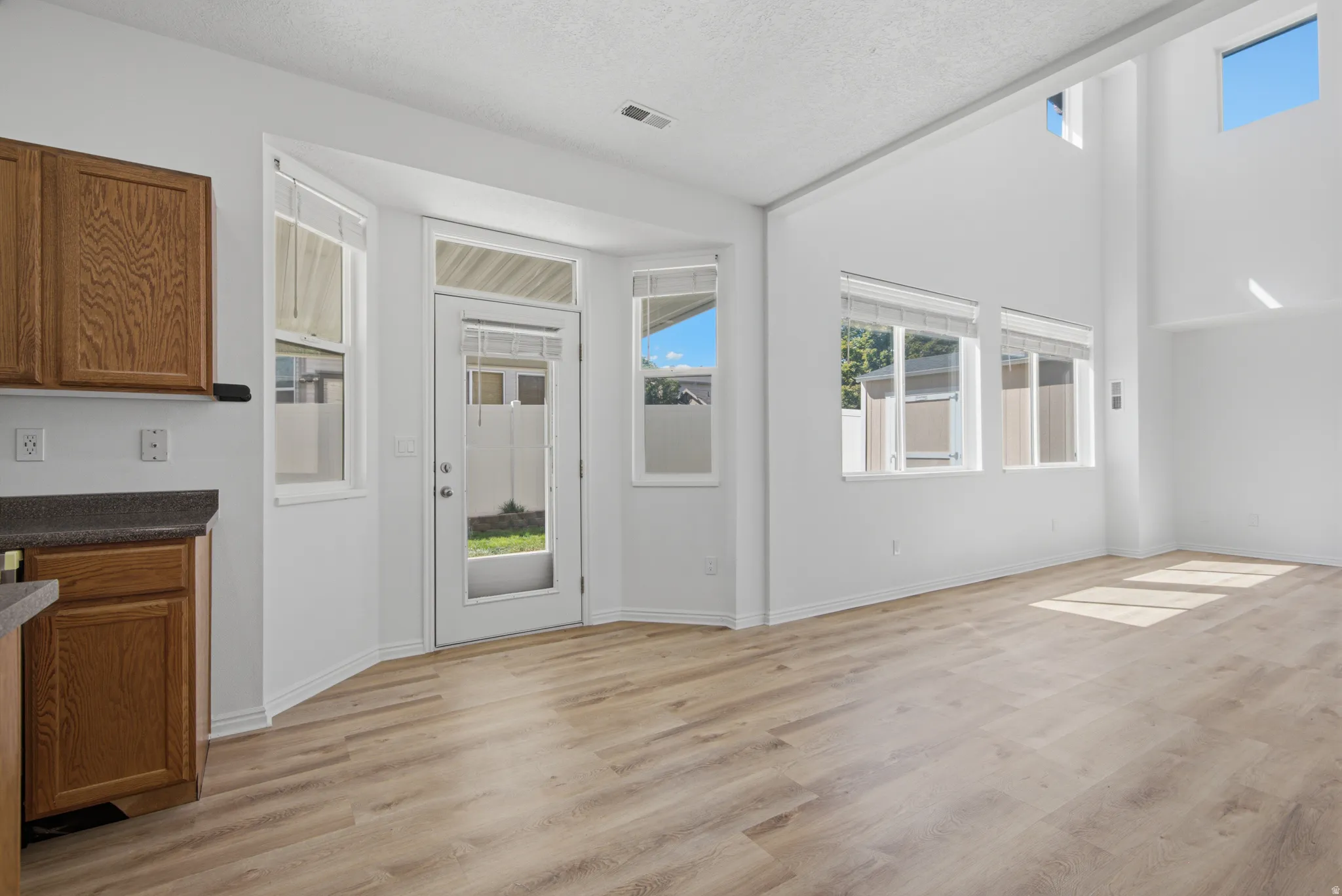 Vaulted ceiling with high windows, wood-finish flooring, white walls, kitchen cabinetry with dark countertops, and a glass-paneled door