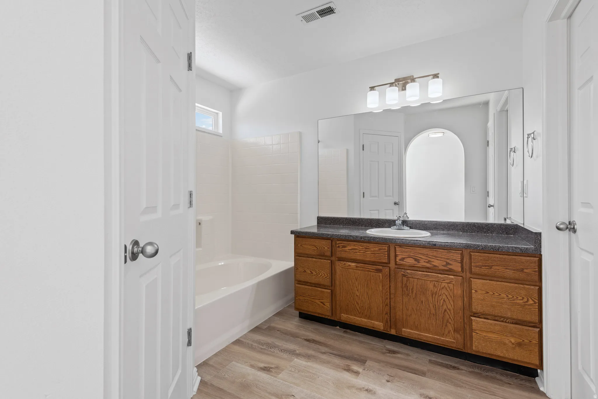Spacious bathroom featuring a full-width vanity with wood-finish cabinetry, a large integrated mirror, and a white soaking tub with tiled surround