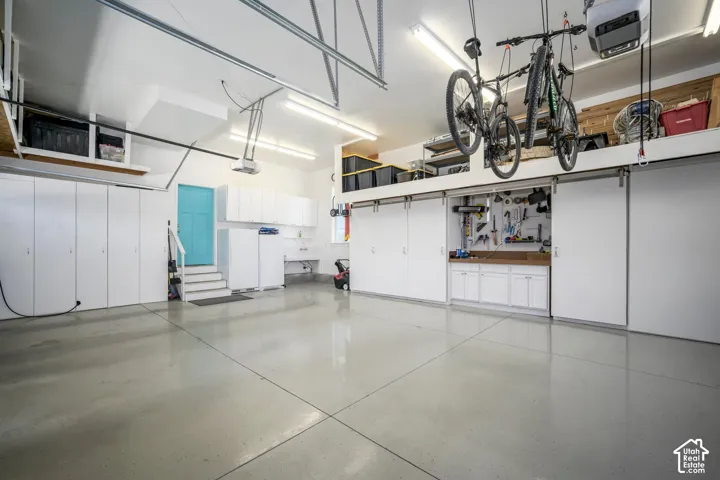 Spacious garage with light grey flooring, extensive built-in white cabinetry, a dedicated workbench area, and overhead storage shelving
