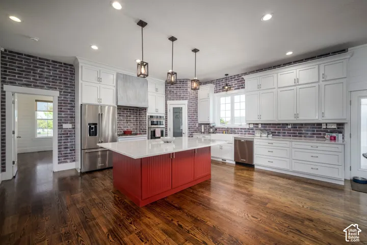 Spacious kitchen featuring rich wood-finish flooring, white cabinetry, a red island with a white countertop, stainless steel appliances, and exposed brick walls
