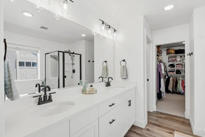 Dual vanity with white cabinetry and integrated sinks, oversized mirror, walk-in shower with black-framed glass enclosure, wood-finish flooring, and recessed lighting