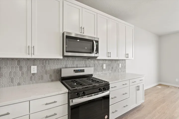 **DISCLAIMER** Some upgrades are not photographed. White shaker-style cabinetry with brushed nickel hardware, light-toned countertops, full-height gray picket tile backsplash, stainless steel microwave, and a black gas range