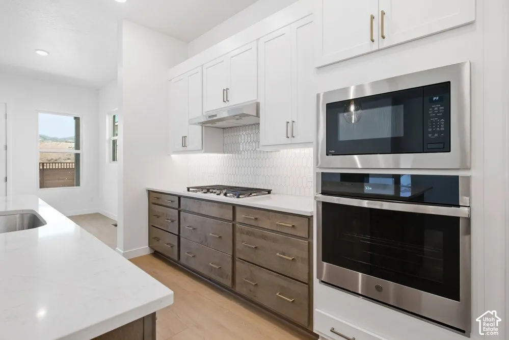 Kitchen featuring white upper cabinetry, lower cabinets with a wood finish, light-toned countertops, a built-in stainless steel double oven and microwave, and a geometric tile backsplash