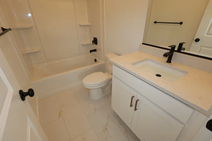 White tile flooring, integrated shower/tub unit, white vanity with gold-tone hardware, light-toned countertop with integrated sink, and matte black fixtures