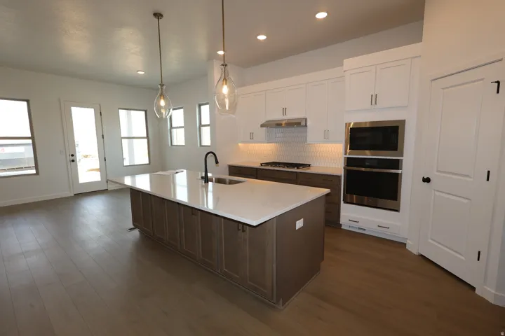 Modern kitchen featuring an expansive center island with an undermount sink and a matte black faucet, wood-finish flooring, recessed lighting, and stainless steel built-in appliances