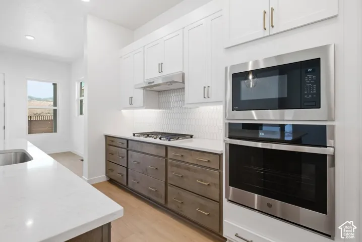 Kitchen featuring white upper cabinetry, lower cabinets with a wood finish, light-toned countertops, a built-in stainless steel double oven and microwave, and a geometric tile backsplash