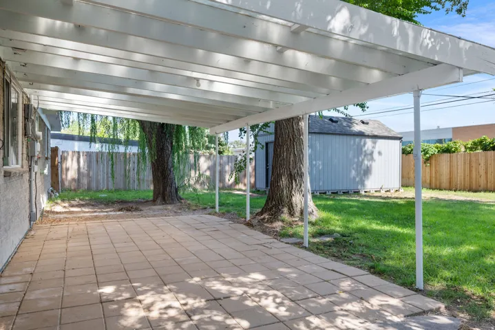 Covered patio with paver flooring, expansive lawn, mature trees, and a storage shed