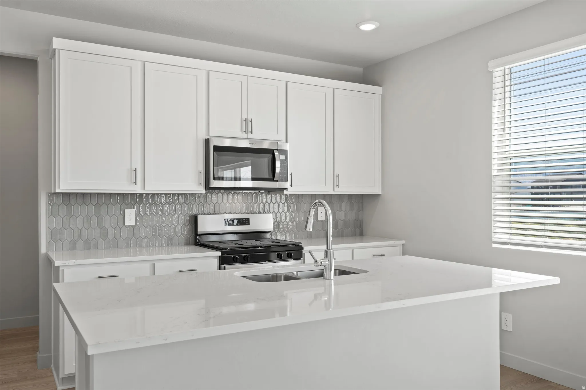 White shaker-style cabinetry, stainless steel appliances, light-toned countertops, tile backsplash, and a kitchen island with an integrated sink