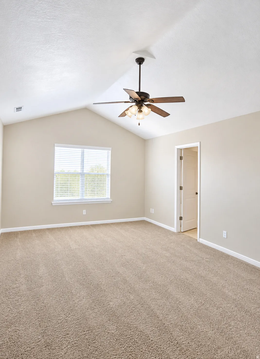 Primary suite with vaulted ceiling, ceiling fan light fixture, neutral carpeting, and window with blinds