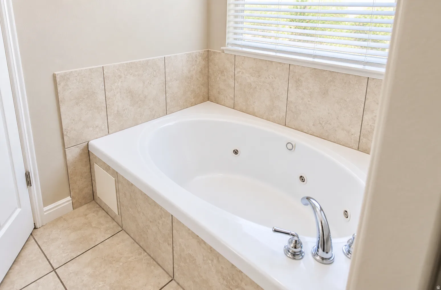 Built-in jetted tub with chrome fixtures, surrounded by neutral-toned wall and floor tiling