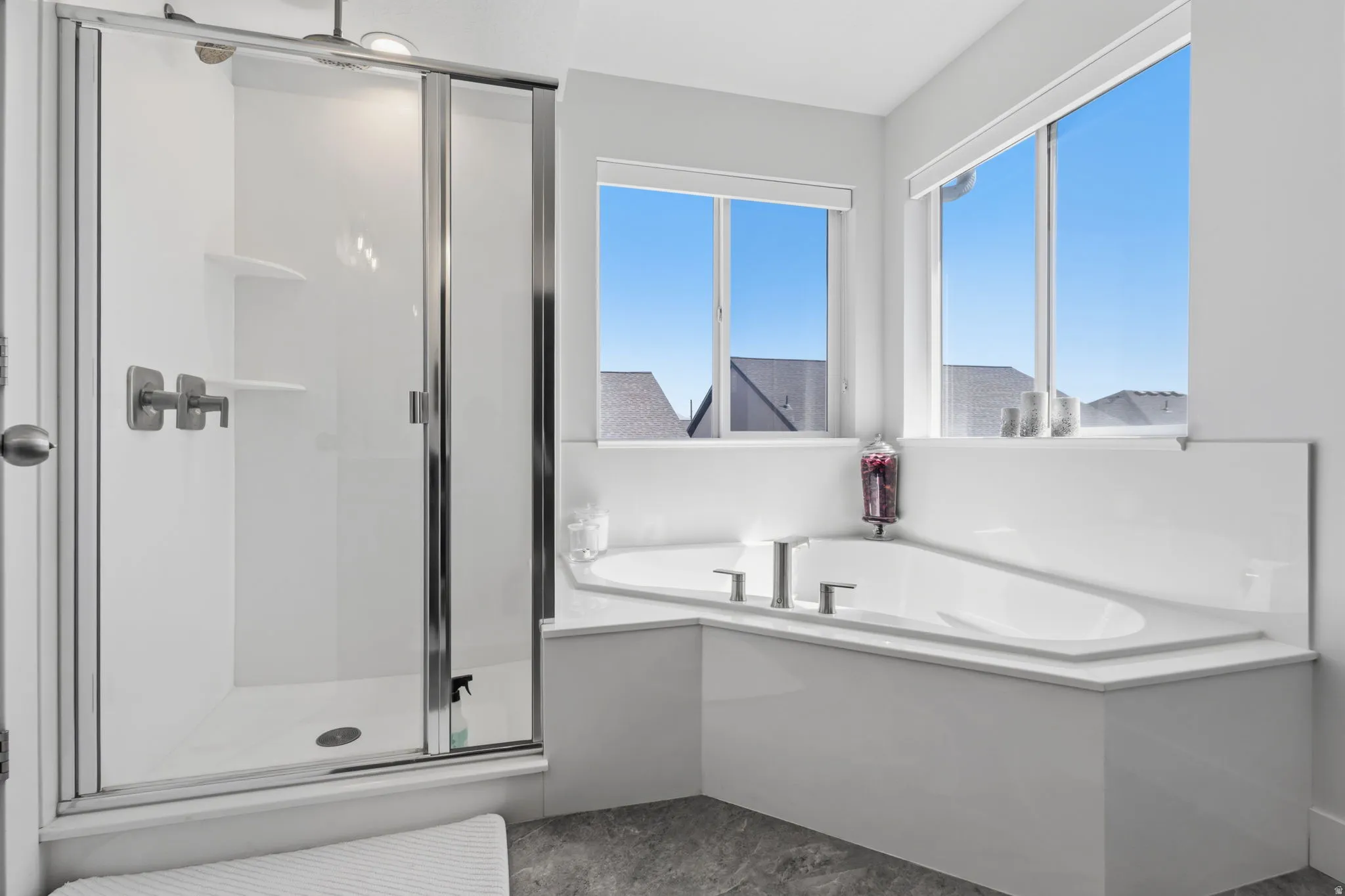 Built-in soaking tub with deck-mounted fixtures, separate shower with glass enclosure, dual windows, and light gray flooring