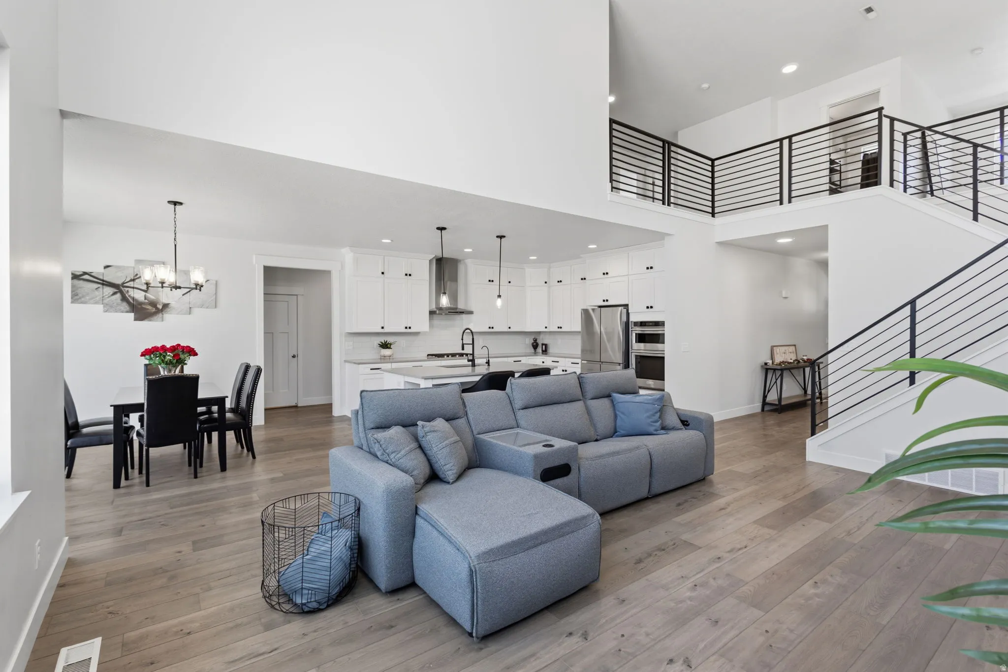 Open-concept living space featuring wood-finish flooring, a two-story ceiling, a contemporary kitchen with white cabinetry, and a staircase with sleek horizontal railings