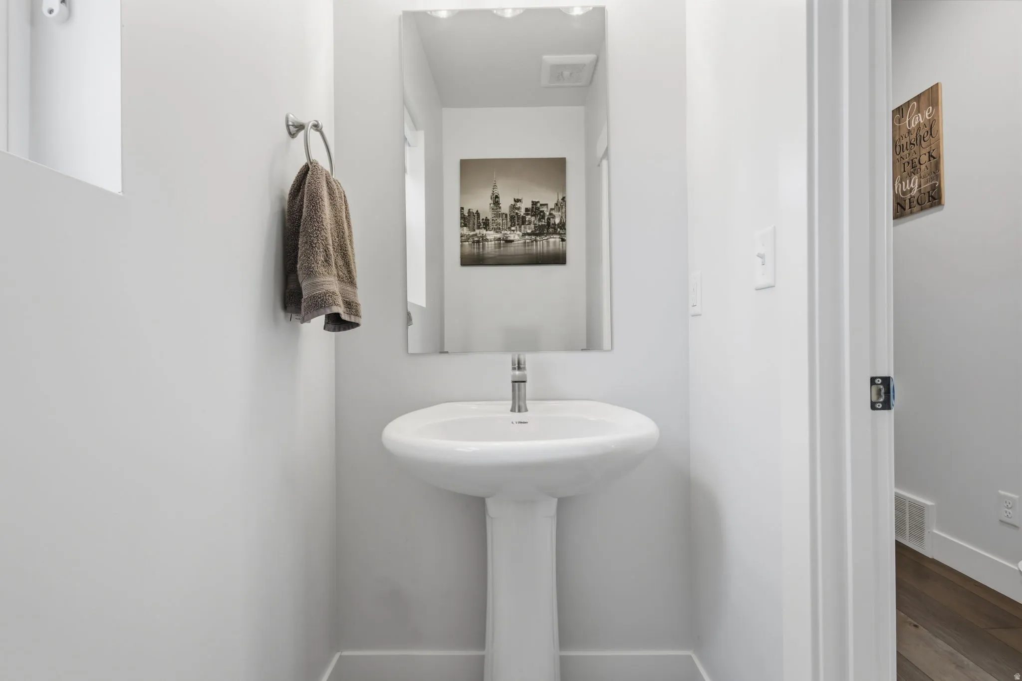 Pedestal sink with a polished chrome faucet, rectangular mirror with overhead lighting, and hardwood-finish flooring visible through the doorway