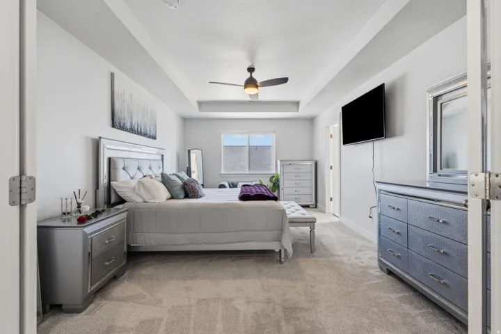 Tray ceiling with a ceiling fan, wall-mounted television, and light-colored carpeting