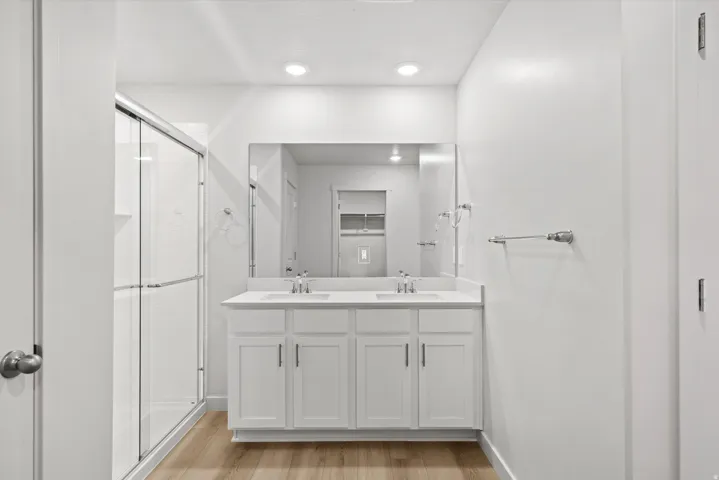 Double vanity with white cabinetry and integrated sinks, a glass-enclosed shower, wood-finish flooring, and recessed lighting