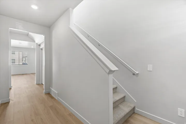 Carpeted staircase with white handrail, leading to a hallway with wood-finish flooring and recessed lighting