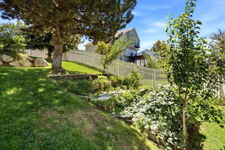 Tiered landscaping with established trees, natural rock retaining walls, a white picket fence, and distant architectural elements featuring a balcony
