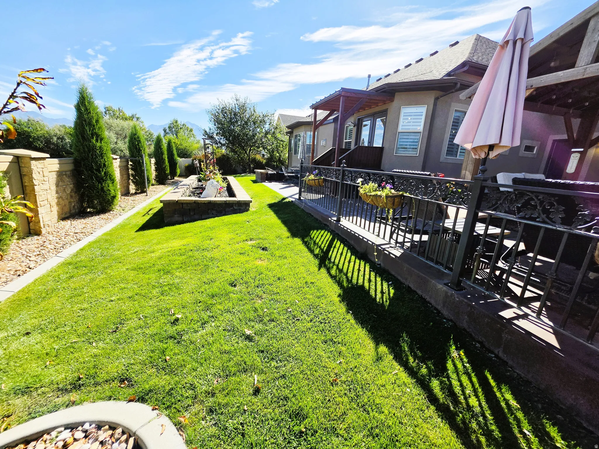 Manicured lawn, elevated patio with decorative railing, stone-clad retaining wall, mature perimeter trees, and built-in planters
