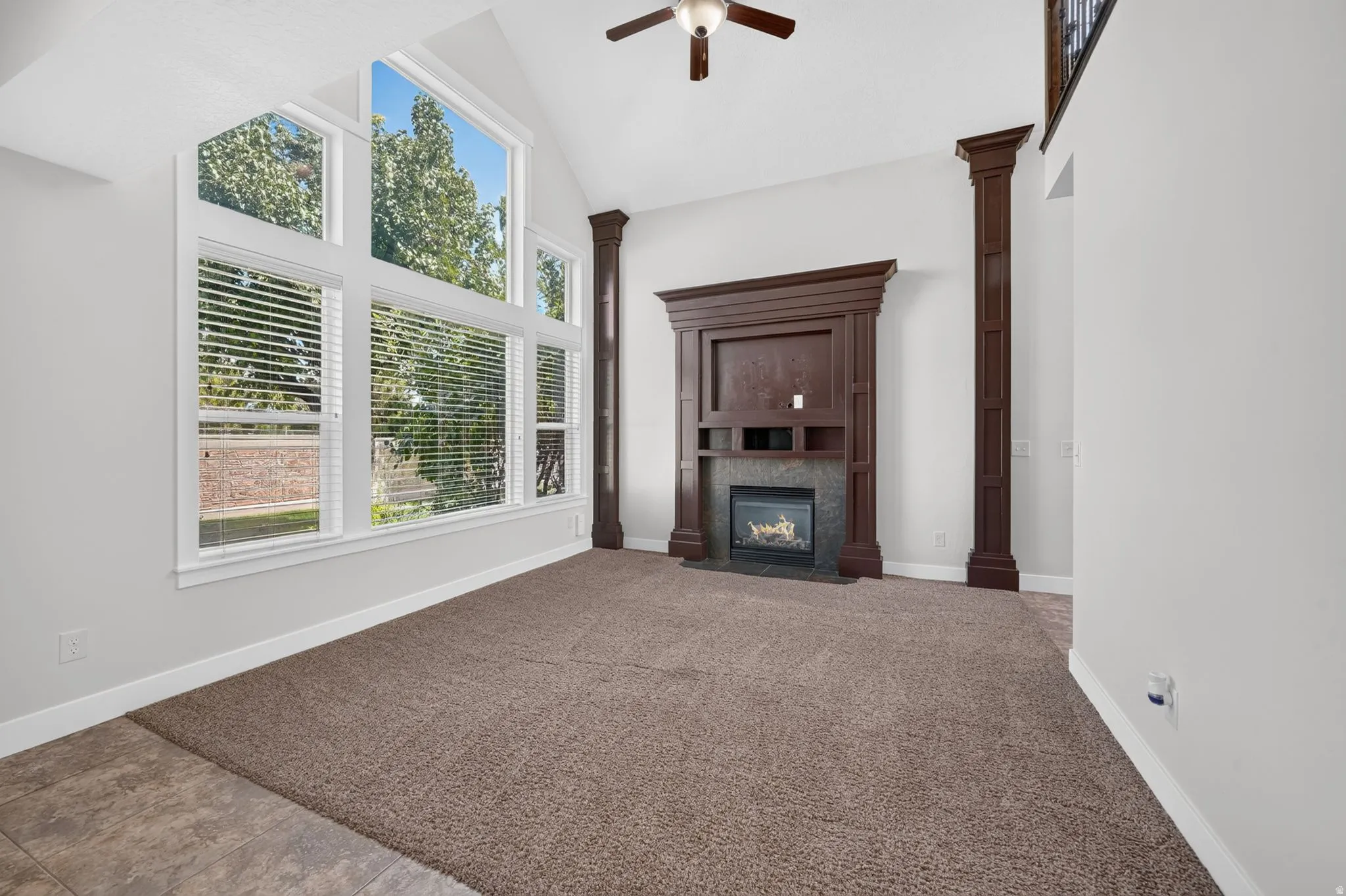 Vaulted ceiling, large windows with blinds, built-in fireplace with dark wood surround, dark wood decorative columns, and a ceiling fan