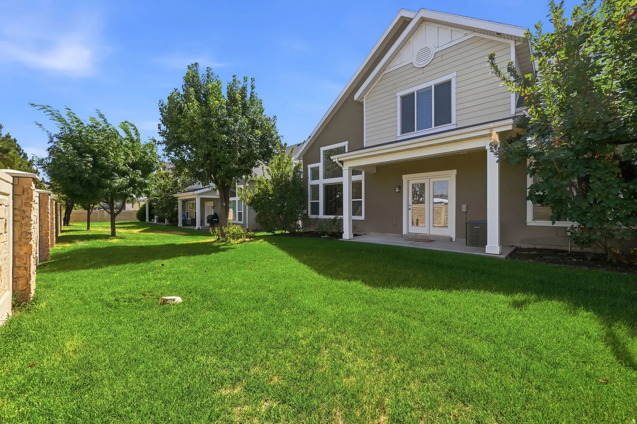Expansive green lawn, mature trees, stone-finish privacy wall, covered patio, and dual glass French doors