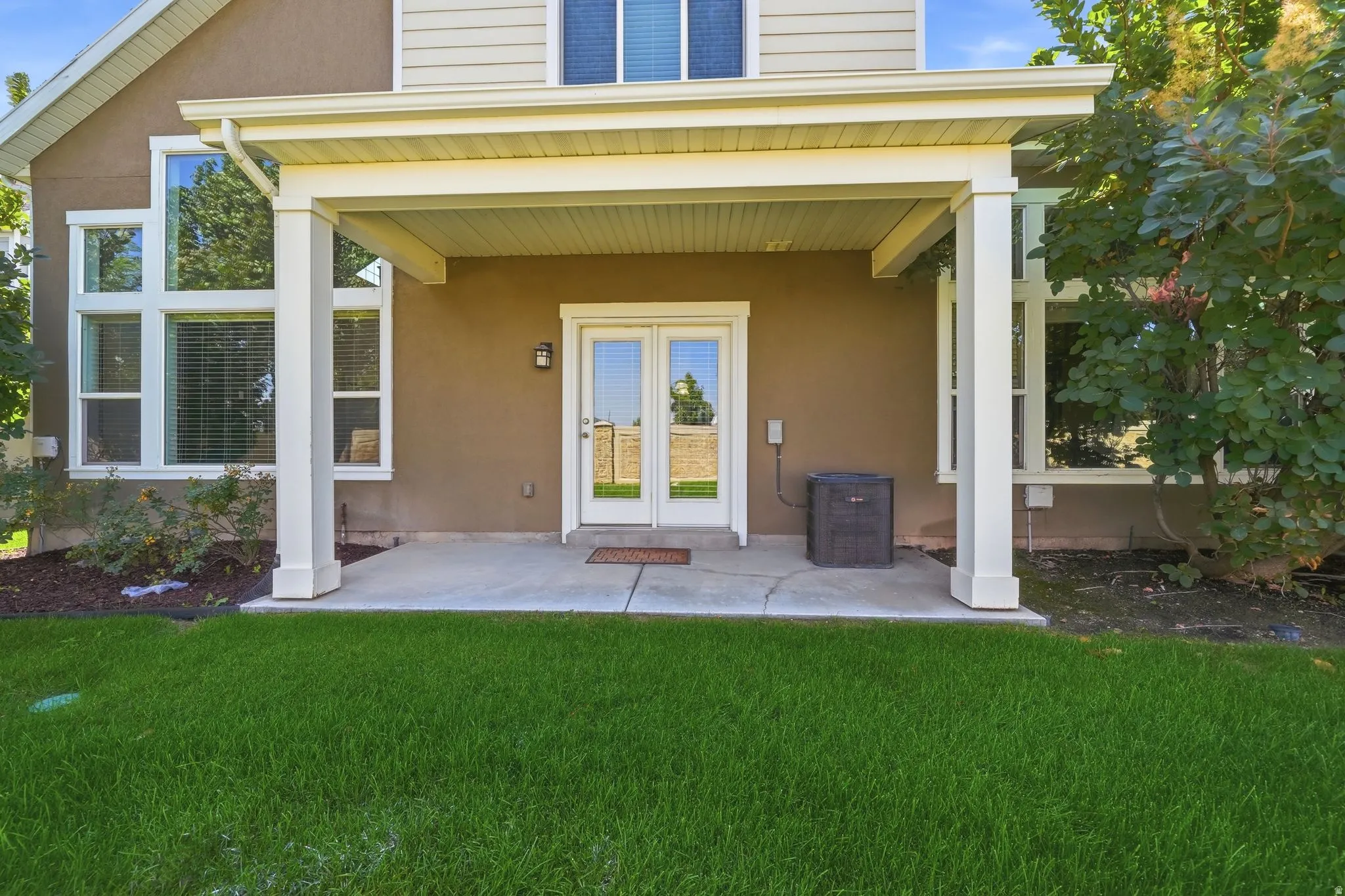 Covered patio with concrete flooring, framed by white columns, featuring French doors with transom windows, and expansive green lawn