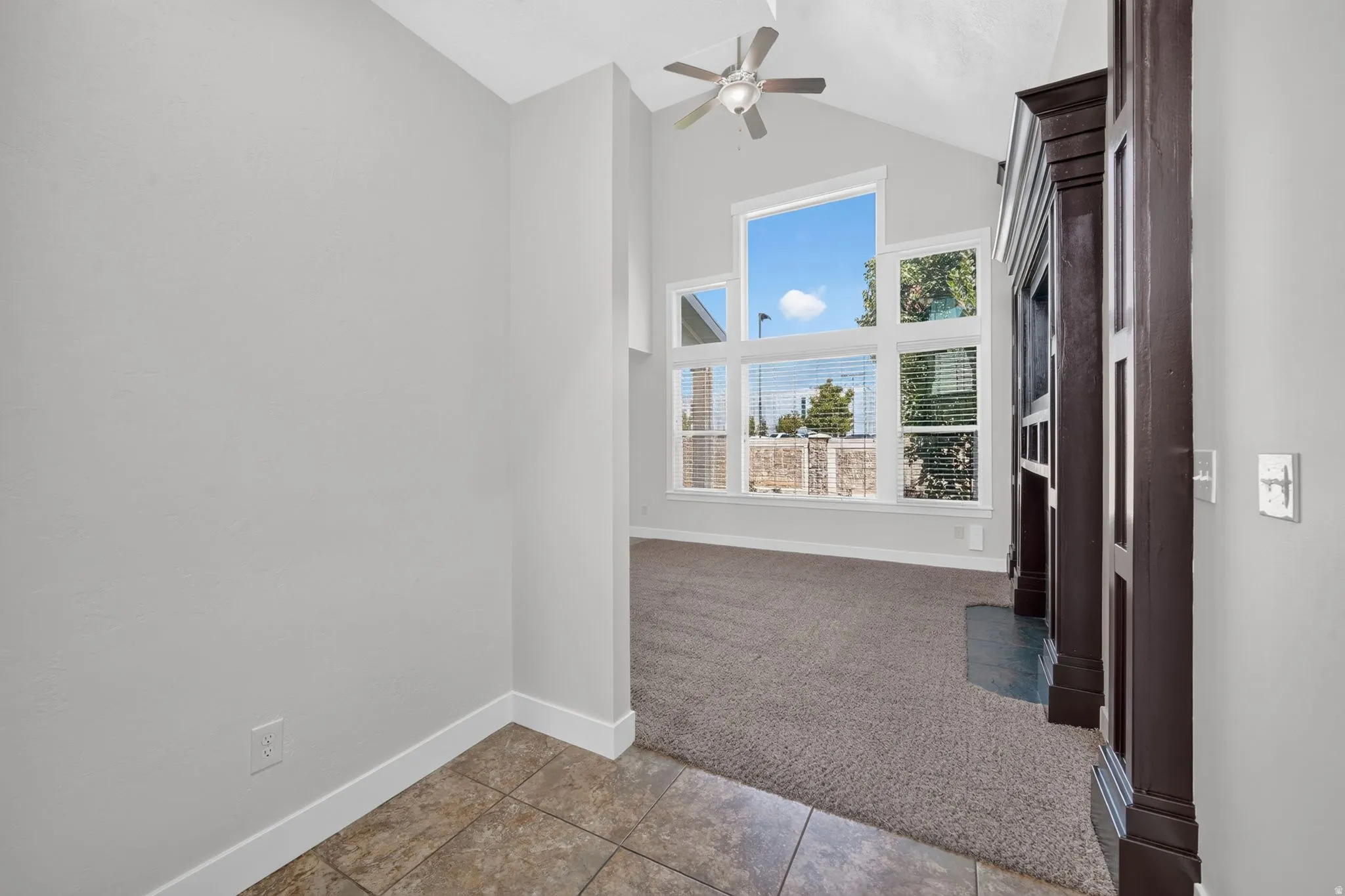 Vaulted ceiling, ceiling fan, large windows, dark wood-tone fireplace surround, and tile flooring