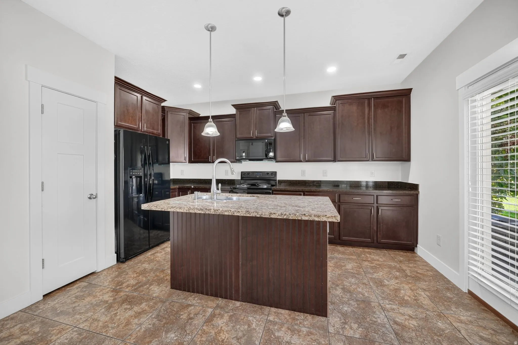 Kitchen featuring dark wood-finish cabinetry, black appliances, a central island with a stone-finish countertop, and pendant lighting