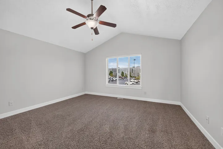 Vaulted ceiling, ceiling fan with light fixture, large window with blinds, and neutral-toned carpeting