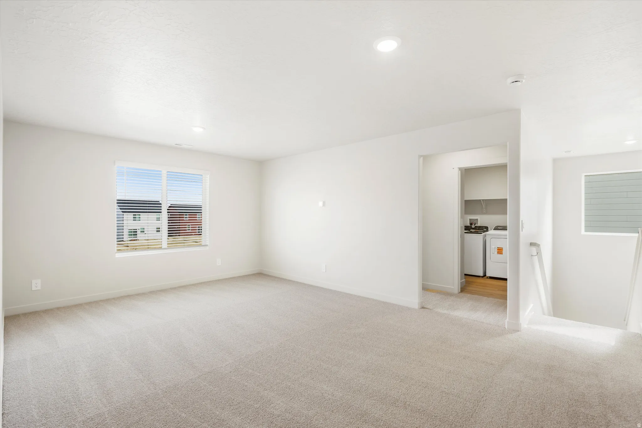 Carpeted living area with recessed lighting, large window with blinds, and access to a laundry space featuring wood-finish flooring and built-in shelving